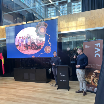Two presenters at a First Australians Capital event, speaking at a podium in front of a large screen displaying an Indigenous community image.